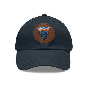 May include: Navy blue baseball cap with a brown leather patch featuring the word "THUNDER" above a stylized bison head in orange and blue. The cap has a curved brim and a classic six-panel design.