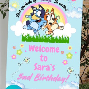 Op de afbeelding: Een lichtblauw verjaardagsbord met de tekenfilmfiguren Bluey en Bingo, een regenboog en ballonnen. Op het bord staat "Welcome to Sara's 5th Birthday!" in roze. Het bord staat op een ezel.
