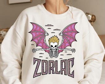 May include: White sweatshirt with a pink and purple cartoon skeleton with wings flying in the clouds. The text "ZORLAC" is printed below the skeleton.