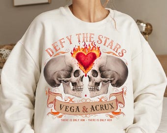 May include: White sweatshirt with a graphic print of two skulls with a heart on fire between them. The text "Defy the Stars" is above the skulls and "Vega & Acrux" is below. The text "There is only him - There is only her" is at the bottom of the graphic.