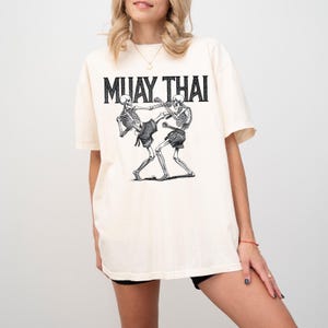 May include: A cream-colored t-shirt with a black graphic of two skeletons practicing Muay Thai. The text "MUAY THAI" is printed in black above the graphic.