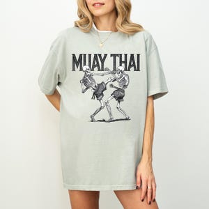 May include: Sage green t-shirt with a graphic of two skeletons practicing Muay Thai. The text "MUAY THAI" is printed in black above the skeletons.