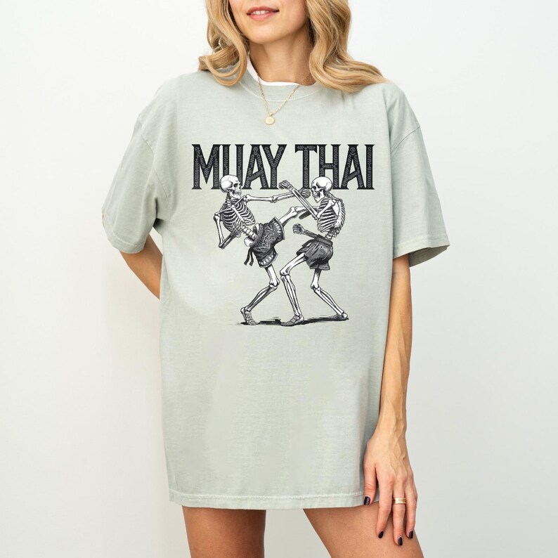 May include: Sage green t-shirt with a graphic of two skeletons practicing Muay Thai. The text "MUAY THAI" is printed in black above the skeletons.