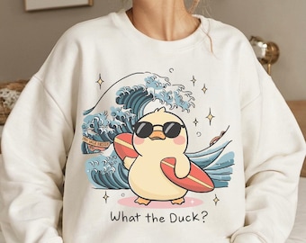 May include: White sweatshirt with a cartoon duck wearing sunglasses and holding a surfboard. The duck is riding a wave with a blue and white design. The text "What the Duck?" is printed below the duck.