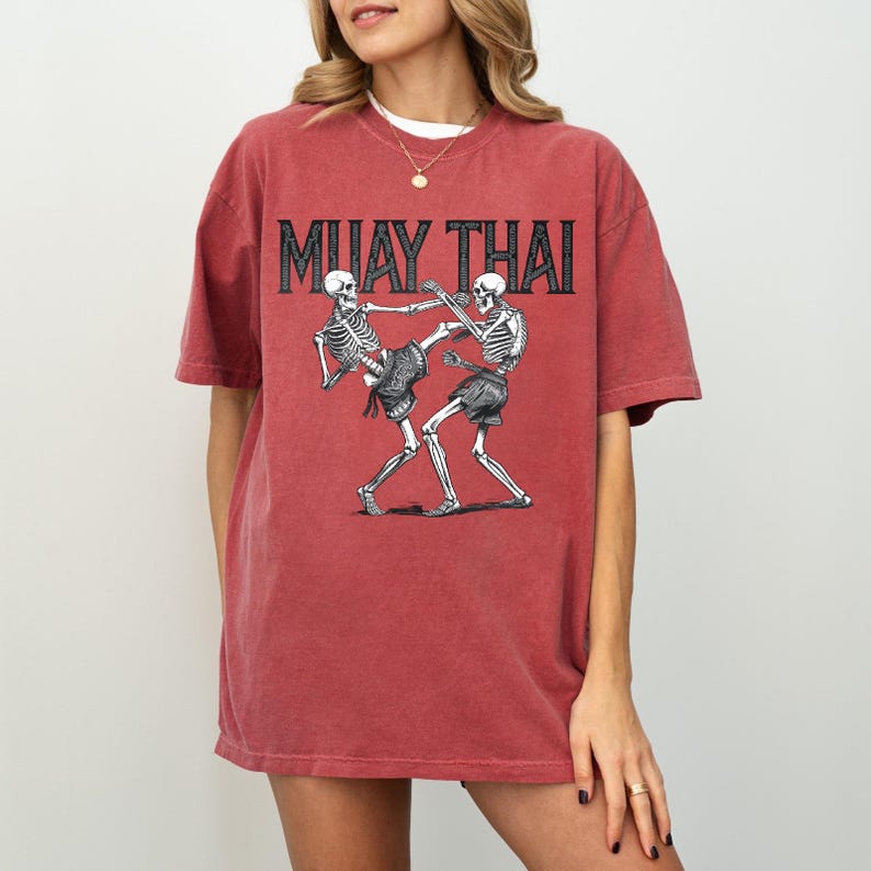 May include: A red, oversized t-shirt with a graphic of two skeletons fighting in a Muay Thai pose. The text "MUAY THAI" is printed in black above the skeletons.