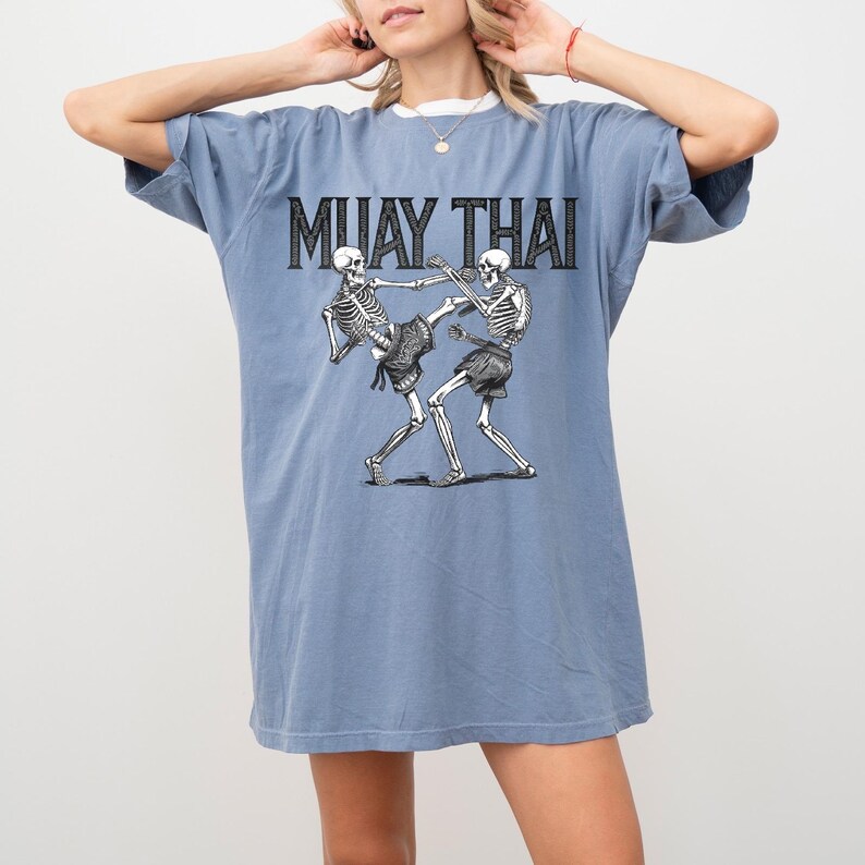 May include: A light blue t-shirt with a graphic of two skeletons practicing Muay Thai. The text "MUAY THAI" is printed in black above the skeletons.