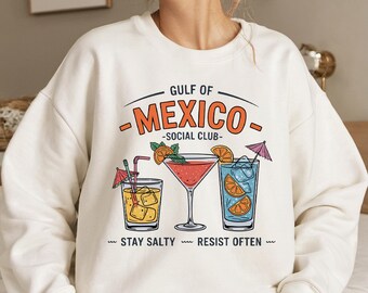 May include: Cream-colored sweatshirt with a graphic of three cocktails and the text "GULF OF MEXICO - SOCIAL CLUB." The cocktails include orange slices and miniature umbrellas. The text "STAY SALTY RESIST OFTEN" is also included.