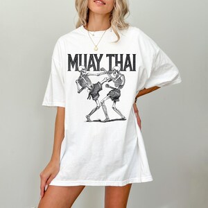 May include: White oversized t-shirt with a graphic of two skeletons practicing Muay Thai. The text "MUAY THAI" is printed above the graphic.