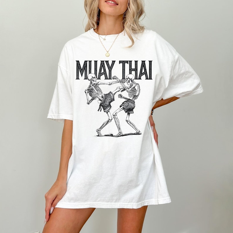 May include: White oversized t-shirt with a graphic of two skeletons practicing Muay Thai. The text "MUAY THAI" is printed above the graphic.