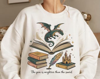 May include: Cream-colored sweatshirt featuring a dragon with open wings above an open book. Below are stacks of books, a quill, and a castle. The text "The pen is mightier than the sword" is printed at the bottom.
