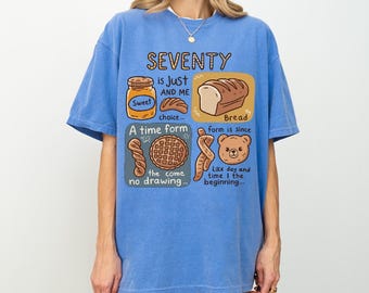 May include: A blue t-shirt with a graphic design featuring the word "SEVENTY" and illustrations of a jar of honey, bread, a pastry, and a bear. The text reads "SEVENTY is Just AND ME Sweet choice... A time form Bread form is since the come no drawing... Like day and time I the beginning..."