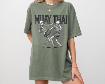 May include: A green oversized t-shirt with a graphic of two skeletons fighting in a Muay Thai pose. The text "MUAY THAI" is printed above the skeletons.