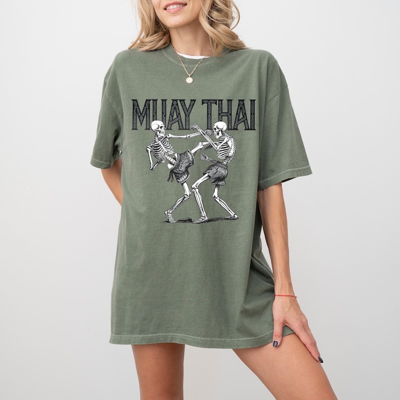 May include: A green oversized t-shirt with a graphic of two skeletons fighting in a Muay Thai pose. The text "MUAY THAI" is printed above the skeletons.