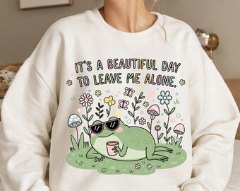 May include: White sweatshirt with a green frog wearing sunglasses and holding a cup. The text on the sweatshirt reads "IT'S A BEAUTIFUL DAY * TO LEAVE ME ALONE. *"