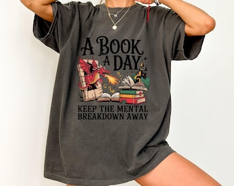 May include: A dark gray t-shirt with a graphic of a dragon breathing fire onto a stack of books. The text "A Book A Day Keep The Mental Breakdown Away" is printed on the shirt.