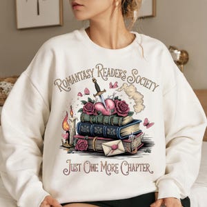 Könnte beinhalten: Weißer Pullover mit einem grafischen Design aus einem Stapel Bücher, einem von einem Dolch durchbohrten Herzen und Schmetterlingen. Der Text "Romantasy Readers Society" und "Just One More Chapter" ist auf dem Pullover aufgedruckt.