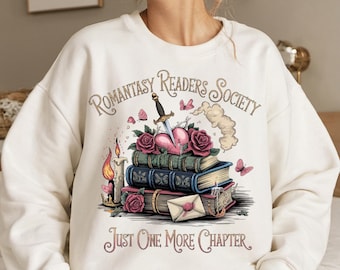 May include: White sweatshirt with a graphic design of a stack of books, a heart pierced by a dagger, and butterflies. The text "Romantasy Readers Society" and "Just One More Chapter" are printed on the sweatshirt.