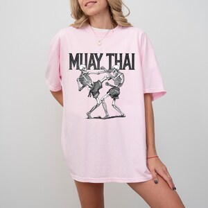 May include: Pink t-shirt with a graphic of two skeletons fighting in a Muay Thai pose. The text "MUAY THAI" is printed in black above the skeletons.