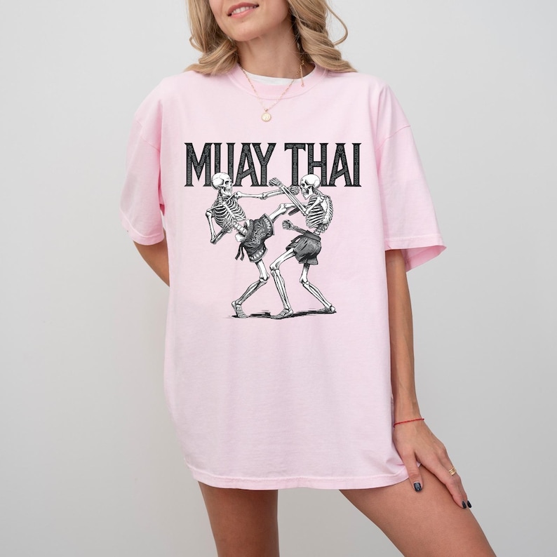May include: Pink t-shirt with a graphic of two skeletons fighting in a Muay Thai pose. The text "MUAY THAI" is printed in black above the skeletons.