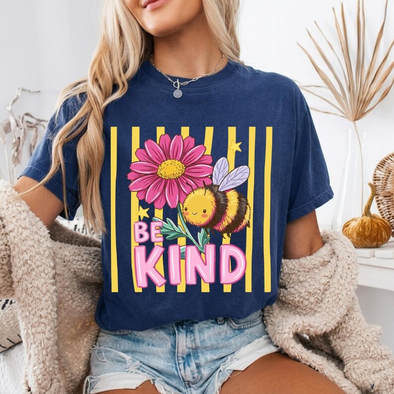 May include: Navy blue t-shirt featuring a graphic of a pink flower, a cartoon bee, and the words "BE KIND" in pink letters. The design is set against a background of yellow vertical stripes.