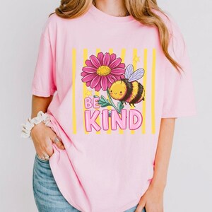 May include: Pink t-shirt featuring a cartoon bee and a pink flower with yellow stripes. The text "BE KIND" is displayed in pink letters. The shirt is a soft, comfortable material.