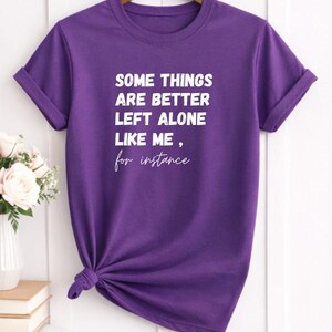 May include: A purple t-shirt with the text "SOME THINGS ARE BETTER LEFT ALONE LIKE ME, for instance" in white. The shirt is knotted at the waist and hangs on a wooden hanger. The sleeves are cuffed.