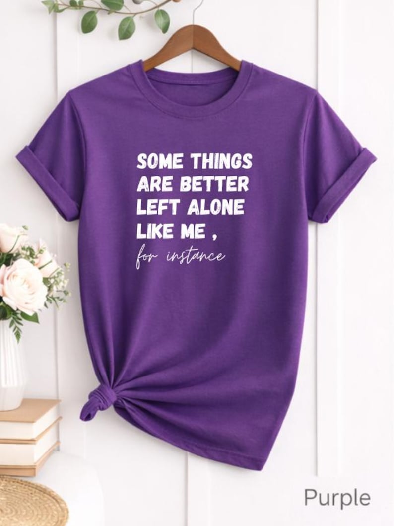 May include: A purple t-shirt with the text "SOME THINGS ARE BETTER LEFT ALONE LIKE ME, for instance" in white. The shirt is knotted at the waist and hangs on a wooden hanger. The sleeves are cuffed.