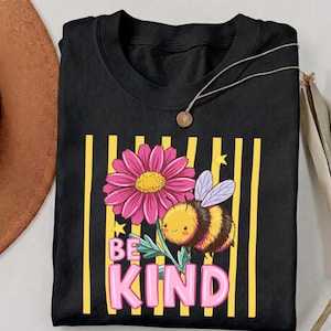 May include: Black t-shirt featuring a pink daisy, a cartoon bee, and the words "BE KIND" in pink letters. The design is set against a background of yellow vertical stripes. A gold necklace is draped over the shirt.