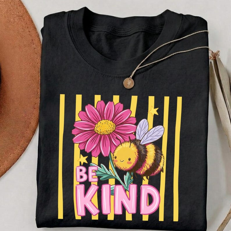 May include: Black t-shirt featuring a pink daisy, a cartoon bee, and the words "BE KIND" in pink letters. The design is set against a background of yellow vertical stripes. A gold necklace is draped over the shirt.