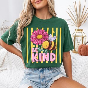 May include: Green t-shirt with a graphic of a pink flower, a smiling bee, and the words "BE KIND" in pink. The design is set against yellow vertical stripes.