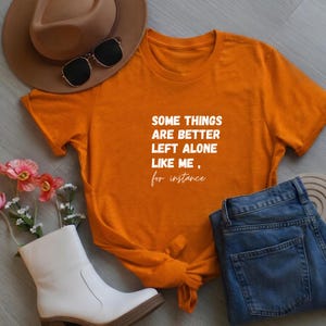May include: Orange t-shirt with the text "SOME THINGS ARE BETTER LEFT ALONE LIKE ME, for instance." The shirt is paired with blue jeans, a brown hat, sunglasses, white boots, and flowers. A casual, everyday outfit.