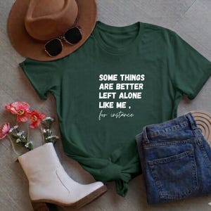 May include: Dark green t-shirt with the text "SOME THINGS ARE BETTER LEFT ALONE LIKE ME, for instance" in white. The shirt is paired with blue jeans, a brown hat, sunglasses, and white boots. Flowers are also visible.
