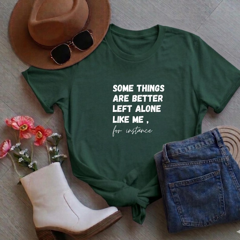 May include: Dark green t-shirt with the text "SOME THINGS ARE BETTER LEFT ALONE LIKE ME, for instance" in white. The shirt is paired with blue jeans, a brown hat, sunglasses, and white boots. Flowers are also visible.