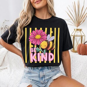 May include: Black t-shirt with a graphic of a pink daisy, a smiling bee, and the words "BE KIND" in pink. The design is set against yellow vertical stripes.
