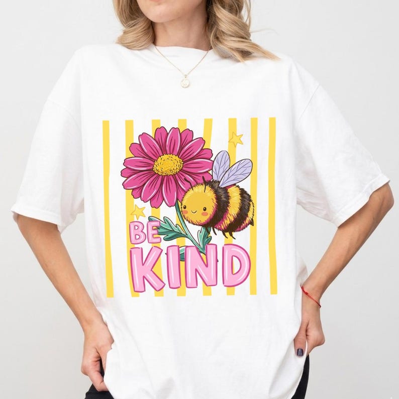 May include: White t-shirt featuring a cartoon bee and a pink flower with the words "BE KIND" in pink letters. The design includes yellow vertical stripes and small stars. The shirt is a casual, everyday wear item.