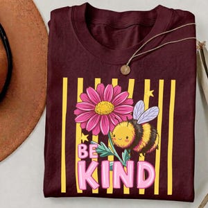 May include: Burgundy t-shirt with a floral and bee graphic. The design includes a pink flower, a cartoon bee, and the words "BE KIND" in pink letters. Yellow vertical stripes are behind the graphic.
