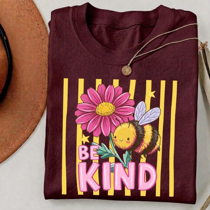 May include: Burgundy t-shirt with a floral and bee graphic. The design includes a pink flower, a cartoon bee, and the words "BE KIND" in pink letters. Yellow vertical stripes are behind the graphic.