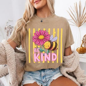 May include: Tan t-shirt with a graphic of a pink flower, a cartoon bee, and the words "BE KIND" in pink. The design is set against a yellow striped background. The shirt is short-sleeved.