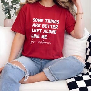 May include: A red t-shirt with the text "SOME THINGS ARE BETTER LEFT ALONE LIKE ME, for instance" in white. The shirt is paired with light blue ripped jeans. The person is sitting on a white couch.