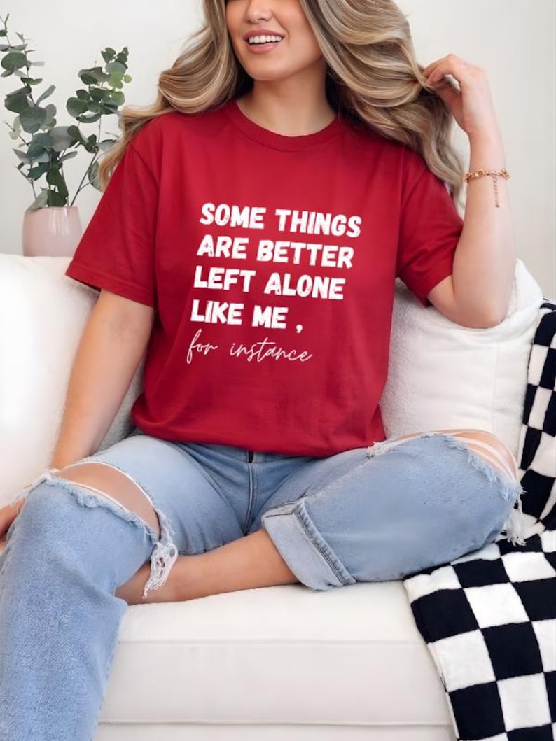 May include: A red t-shirt with the text "SOME THINGS ARE BETTER LEFT ALONE LIKE ME, for instance" in white. The shirt is paired with light blue ripped jeans. The person is sitting on a white couch.