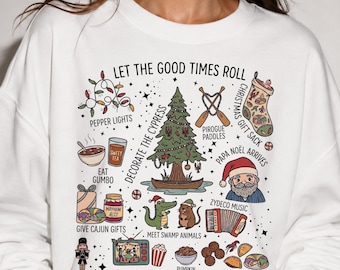 May include: A white crewneck sweatshirt featuring a colourful graphic print with the text LET THE GOOD TIMES ROLL. The design includes illustrations of a cypress tree, a Santa figure, swamp animals, food items, and various holiday-themed icons.