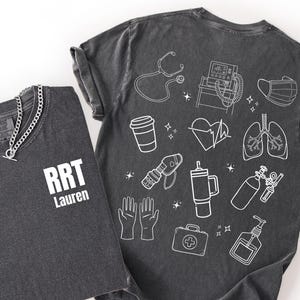 Puede incluir: Dos camisetas de color gris oscuro. Una camiseta tiene el texto "RRT Lauren" y un collar de cadena plateada. La otra camiseta tiene dibujos lineales blancos de equipos médicos, incluyendo un estetoscopio, pulmones y un corazón. La palabra "Pepper" está en la parte inferior.