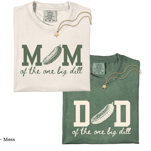 May include: An ivory t-shirt with dark green text reading MOM of the one big dill and a moss green t-shirt with ivory text reading DAD of the one big dill. Both shirts feature a pickle graphic replacing the letter O and a gold star necklace.