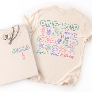 May include: Two cream-colored t-shirts. The folded shirt features the text MAMA and a candle graphic. The unfolded shirt displays the text ONE-DER THE SEA, Sophia's First Birthday, and various colorful sea life illustrations including a crab and starfish.