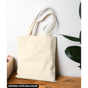 Puede incluir: Un bolso tote de color natural con asas largas se encuentra contra una pared blanca. La bolsa est&aacute; hecha de una tela resistente y est&aacute; vac&iacute;a. El texto "NATURAL TOTE BAG COLOR" est&aacute; en la parte inferior de la imagen. Una cesta y una planta est&aacute;n en el fondo.