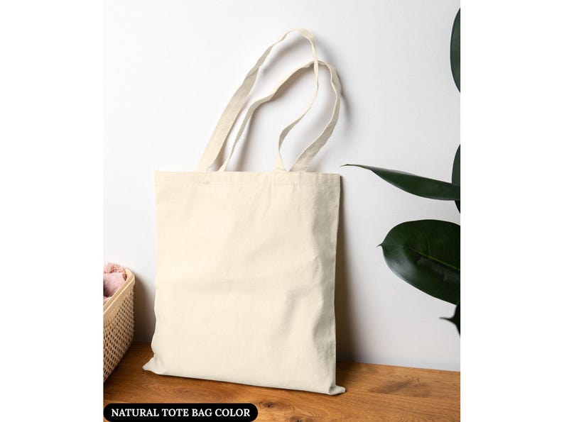 Puede incluir: Un bolso tote de color natural con asas largas se encuentra contra una pared blanca. La bolsa est&aacute; hecha de una tela resistente y est&aacute; vac&iacute;a. El texto "NATURAL TOTE BAG COLOR" est&aacute; en la parte inferior de la imagen. Una cesta y una planta est&aacute;n en el fondo.