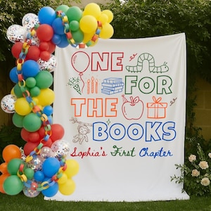 May include: A white fabric backdrop featuring the text ONE FOR THE BOOKS and Sophia's First Chapter with colorful illustrations of a balloon, caterpillar, books, apple, gift, and monkey, accented by a large balloon arch and paper chain garland.