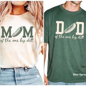 May include: An ivory t-shirt with dark green text reading MOM of the one big dill featuring a pickle graphic, and a blue spruce t-shirt with cream text reading DAD of the one big dill featuring a pickle graphic, both with short sleeves.