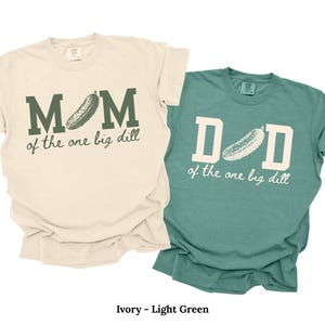 May include: An ivory t-shirt with dark green text reading MOM of the one big dill and a light green t-shirt with cream text reading DAD of the one big dill. Both shirts feature a graphic of a pickle replacing the letter O or A.
