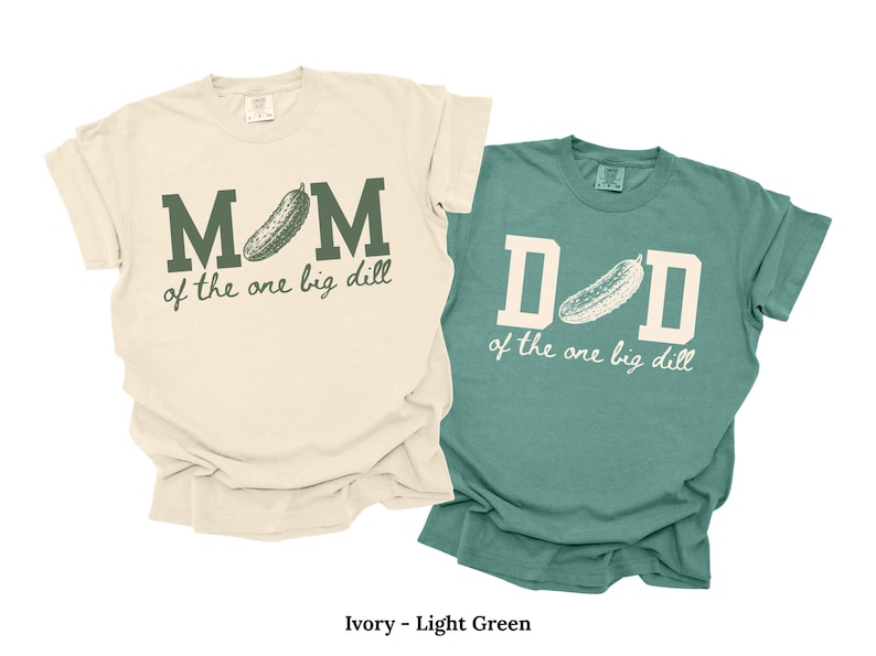 May include: An ivory t-shirt with dark green text reading MOM of the one big dill and a light green t-shirt with cream text reading DAD of the one big dill. Both shirts feature a graphic of a pickle replacing the letter O or A.